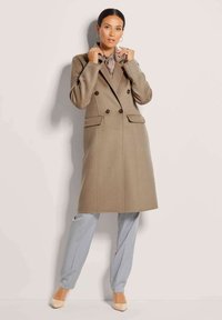 Beige Doppelreiher-Mantel aus strukturiertem Stoff, mit Reverskragen und zwei Fronttaschen. Kombiniert mit hellblauen Hosen und nudefarbenen Absatzschuhen.