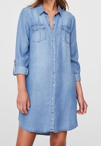 Kvinna iklädd en ljusblå denimskjortklänning med långa ärmar uppvikta och framknäppning.