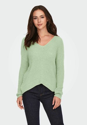 Light green, textured knit sweater with a V-neck and long sleeves, featuring a slightly asymmetrical hem and relaxed fit over dark jeans.