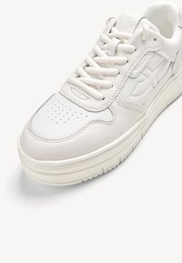 White leather sneaker with textured stitching, circular perforations on the toe, and a chunky sole. Features flat laces and embossed logo accents.