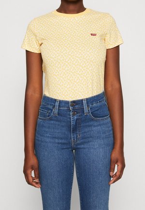 Kobieta w żółtej koszulce z kwiatowym wzorem, z czerwonym logo Levi's oraz wysokim stanem niebieskich dżinsów.