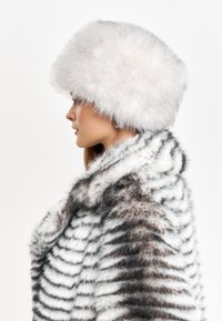 Cappello in finta pelliccia bianco con forma arrotondata, dotato di una fascia spessa e testurizzata. Abbinato a un cappotto in finta pelliccia a strisce grigie e bianche.