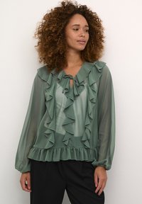 Blusa verde trasparente con dettagli arruffati, collo legato, maniche lunghe e orlo frillato. Tessuto liscio con dettagli a strati.