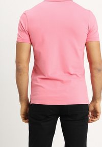 Pánská polo tričko s krátkým rukávem v jednobarevné růžové, vyrobené z hladkého materiálu. Má límec a boční rozparky na lemování, nošeno s černými kalhotami.