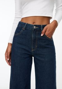 Mujer con jeans de tiro alto azul oscuro, con la mano en el bolsillo y una camiseta larga blanca de corte crop, mostrando el abdomen desnudo, contra un fondo blanco.