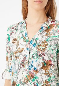 Blusa floral con cuello tipo camisa, que presenta un fondo blanco decorado con coloridos patrones florales en tonos verdes, azules y marrones.