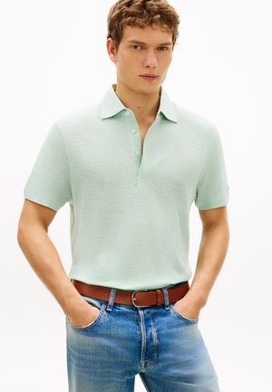 Junger Mann, der ein hellgrünes, kurzärmliges Polohemd trägt, das in blaue Jeans mit braunem Gürtel gesteckt ist, Hände in den Taschen, neutraler Hintergrund.