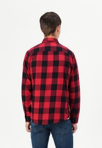 Hombre de espaldas vistiendo una camisa de cuadros rojos y negros de manga larga y jeans azules, frente a un fondo blanco liso.
