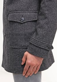 Cappotto di lana grigia con tessuto testurizzato, due tasche frontali con pattine e dettagli in bottone sui polsini. Vista laterale con una mano sul cappotto.
