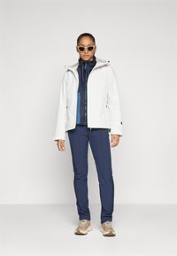Witte waterdichte jas met een kap, gedragen over een marineblauwe fleece. Gecombineerd met marineblauwe broek en beige sneakers, geaccessorieerd met een zonnebril.