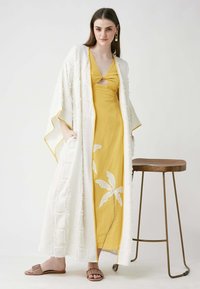 White textured long kimono with fringed edges over a yellow dress featuring a cutout and white leaf applique. Tan slide sandals are worn.