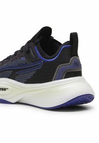 Fekete sportcipő, textúrált hálós felsőrésszel, lila kiegészítőkkel, párnázott nyakkal és vastag fehér középtalppal, amelyen kapaszkodó minta található.