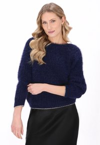 Donkerblauwe fuzzy trui met een wijd decolleté en driekwartmouwen; zilveren accenten aan de onderrand en mouwen. Zwarte rok eronder.
