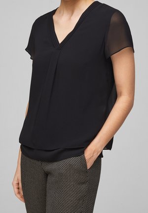 Mujer vistiendo una blusa negra de manga corta con escote en V, mangas transparentes y pantalones entallados estampados, con una mano en el bolsillo.