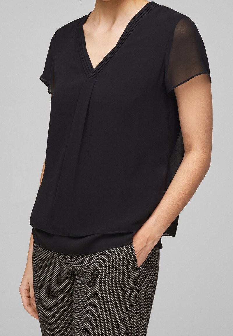 Femme portant un chemisier noir à col en V manches courtes avec des manches transparentes et un pantalon slim à motifs, une main dans la poche.