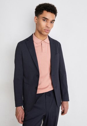 Blazer blu navy su una polo rosa chiaro, entrambi con texture morbide. Il blazer ha un design a un petto e tasche frontali.
