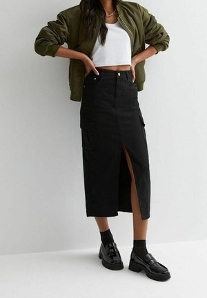 Femme portant un crop top blanc, une veste bomber verte, une jupe midi noire avec une fente devant et des poches cargo, ainsi que des mocassins noirs avec des chaussettes.