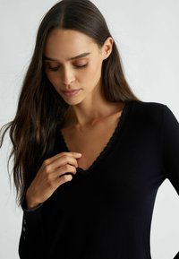 Zwarte V-halstrui met een getextureerde, geschulpte halslijn en lange mouwen met knoopaccenten op de manchetten. Zachte stofafwerking.