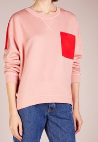 Sudadera de color rosa claro con un bolsillo rojo, cuello redondo y detalles cosidos. Textura suave, ajuste holgado y mangas largas.