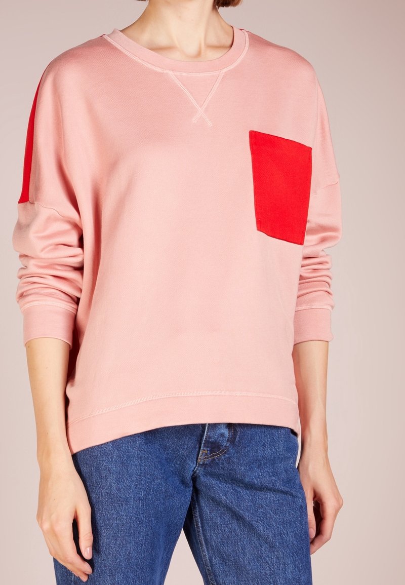 Sudadera de color rosa claro con un bolsillo rojo, cuello redondo y detalles cosidos. Textura suave, ajuste holgado y mangas largas.