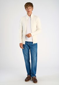 Vit stickad zip-up kofta med diamantmönster, bärs över en enkel vit t-shirt, matchad med blå jeans och bruna snörskor.