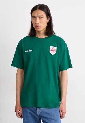 Grøn bomulds t-shirt med rund hals, korte ærmer og et broderet badge på venstre bryst. Har en hvid logo på forsiden.