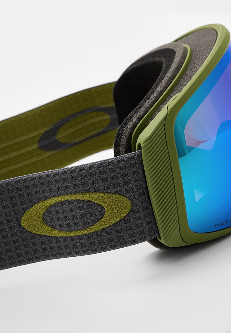 Zielone gogle narciarskie z niebieskim szkłem, teksturowany szary pasek z okrągłym logo. Oprawka ma żłobioną strukturę zapewniającą chwyt.