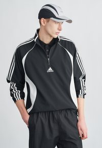 Μαύρο αθλητικό φούτερ με φερμουάρ και λευκές λεπτομέρειες, με τρεις ρίγες στα μανίκια. Φορεμένο με μαύρο αθλητικό παντελόνι και μαύρο/γκρι καπέλο.