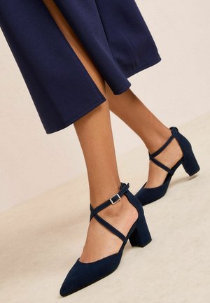 Femeie purtând pantofi cu toc bloc albastru închis, cu barete încrucișate și catarame la gleznă, asortați cu pantaloni largi albastru închis, scurtați.