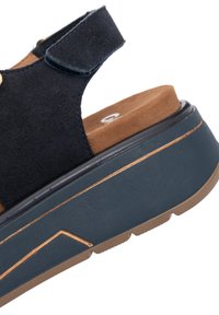 Sandale compensée en daim noir avec bride ajustable à velcro, semelle intérieure marron rembourrée et semelle plateforme bleu marine avec détails beige.