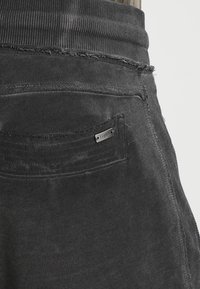 Svarta joggbyxor i ett texturerat material, med ribbat midjeband, slitna detaljer och en bakficka med en metalldekor.