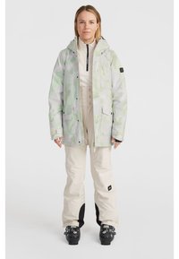 Light green camo jacket with drawstring hood, zippered pockets, and elastic cuffs, paired with tan pants featuring black accents and ski boots.