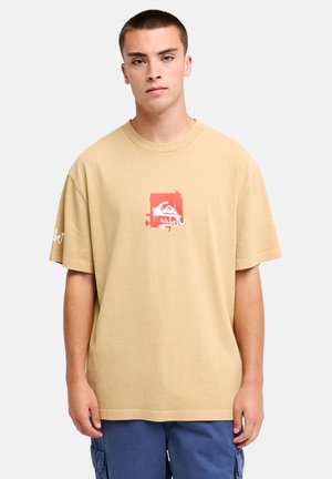 Młody mężczyzna z krótkimi włosami, w luźnym beżowym t-shircie z czerwono-białym nadrukiem i niebieskich szortach cargo, stoi na jednolitym tle.