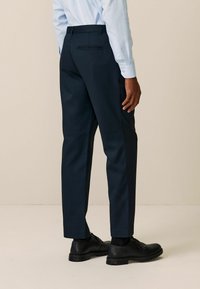 Dunkelnavy Hose mit schmalem Schnitt, mit einer einzelnen Gesäßtasche. Kombiniert mit schwarzen Schuhen, die eine glatte Stoffstruktur zeigen.
