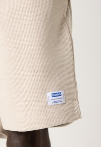 Shorts beige texturés avec un design à côtes. Présente une étiquette bleu et blanc "HUGO" près de l'ourlet. Fabriqué en matériau léger.