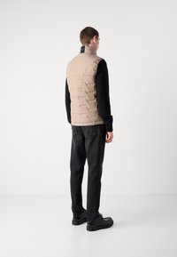 Beige puffervest med høy krage og sydd detaljer, båret over en svart langermet skjorte, kombinert med mørke jeans og svarte sko.