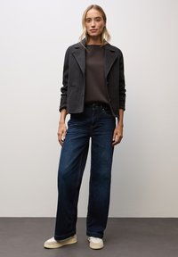 Dunkelgraue Jacke mit breitenrevers, übereinandergetragen über einem braunen Oberteil, kombiniert mit locker sitzenden dunkelblauen Jeans und beigen Sneakers. Glatte Texturen durchgehend.