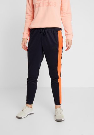 Person iført sorte træningsbukser med klare orange sidelister, ferskenfarvet Adidas sweatshirt og hvide sneakers, står mod en ensfarvet baggrund.