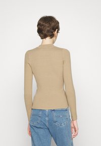 Beige geripptes Langarmoberteil mit hohem Kragen. Kombiniert mit hochgeschnittenen blauen Jeans, die ein Logo-Detail auf der Gesäßtasche zeigen.