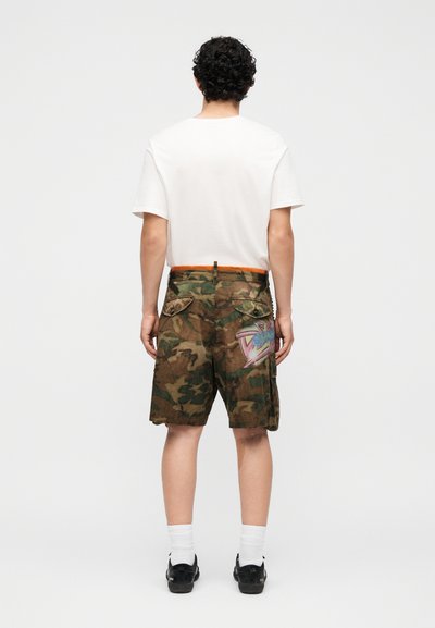 Άνδρας που στέκεται με την πλάτη προς τα πίσω, φοράει λευκό T-shirt, καμουφλάζ cargo σορτς με λεπτομέρειες γκράφιτι, λευκές κάλτσες και μαύρα αθλητικά παπούτσια σε απλό φόντο.