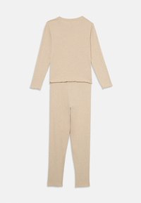 Beige lounge-set av mjukt tyg, innehåller en långärmad topp med rak nederkant och matchande tighta byxor med fransade kanter.