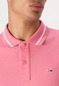 Rosa polo skjorte laget av pustende stoff, med klassisk krage, to knapper og hvite striper. Inkluderer detalj med lite logo.