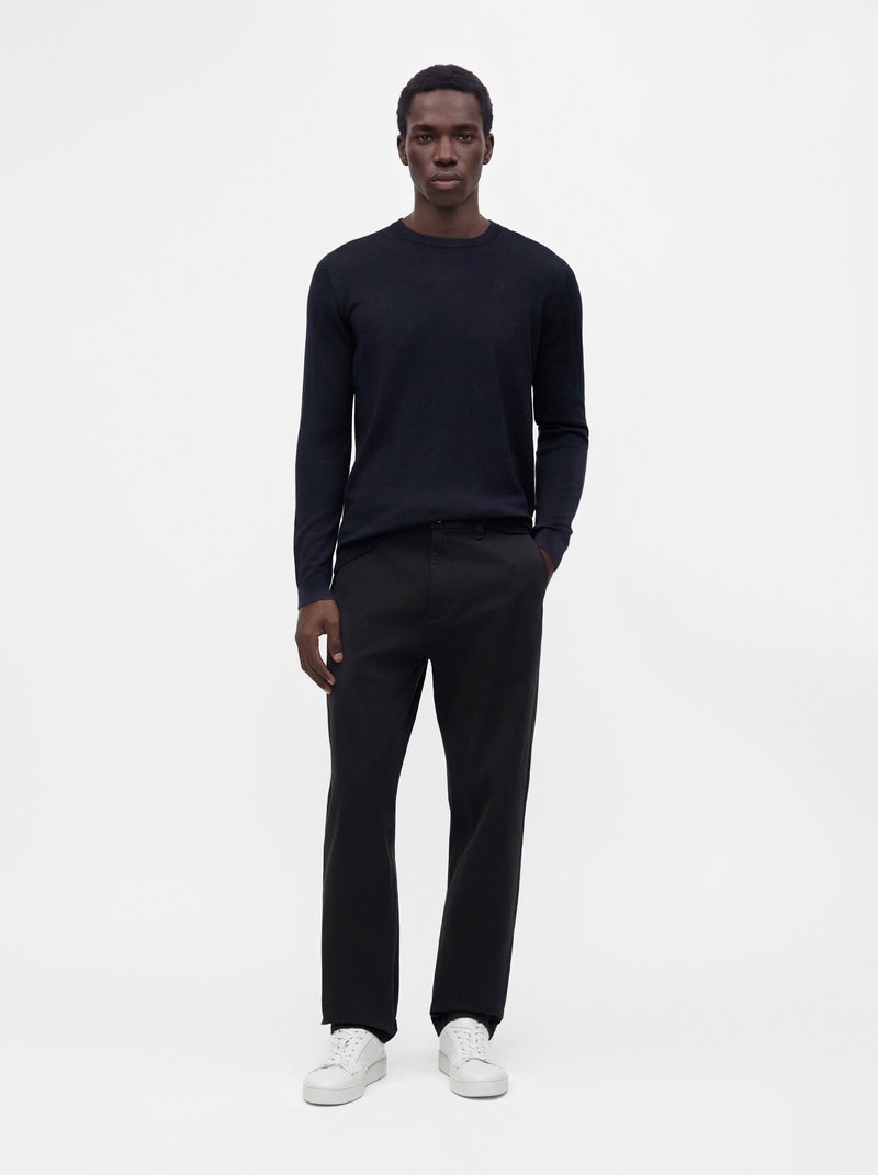 Homme debout portant un pull noir à manches longues, un pantalon noir, des baskets blanches, avec une main dans la poche, sur un fond blanc uni.