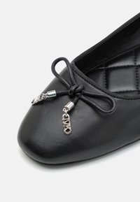 Ballerina di pelle nera con punta tondeggiante, dettaglio con fiocco decorativo e un charm argentato vicino ai lacci. Sola interna con texture visibile.
