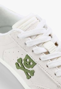 Jasnobeżowe zamszowe sneakersy z białymi sznurowadłami, z zielonym haftem czaszki i przekreślonych kości po bokach. Gładka tekstura, minimalistyczny design.