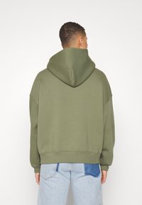Homme portant un sweat à capuche oversized vert olive et un jean bleu clair avec une pièce bleu foncé, vu de dos sur un fond blanc.
