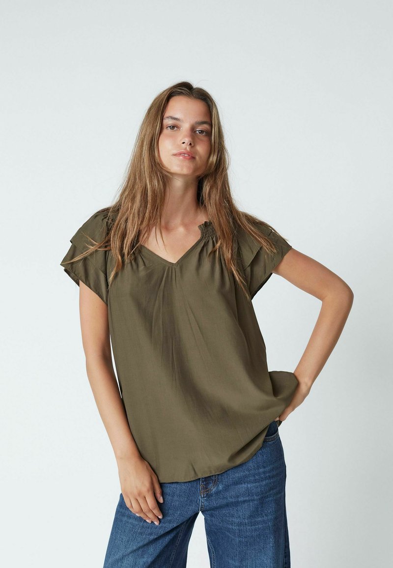 Giovane donna con capelli lunghi che indossa una blusa a maniche corte verde oliva e jeans blu, in piedi con una mano sui fianchi su uno sfondo semplice.