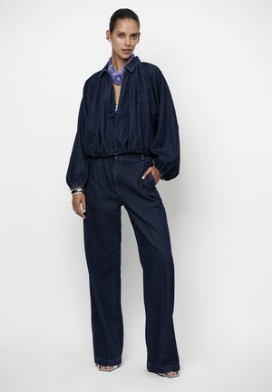 Vrouw draagt donkerblauwe denim wijde broek en een bijpassend volumineus jack met rits, met een paarse nek sjaal en zilveren armbanden.