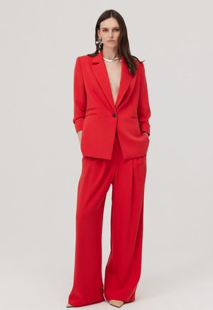 Kobieta ubrana w czerwony, oversize'owy blezer i szerokie spodnie, uzupełniona srebrnym naszyjnikiem oraz dużymi efektownymi kolczykami, stojąca z rękami w kieszeniach.