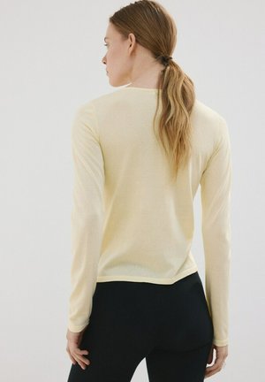 Donna con capelli lunghi legati, che indossa una maglia a maniche lunghe giallo chiaro e pantaloni neri, vista da dietro su uno sfondo neutro.